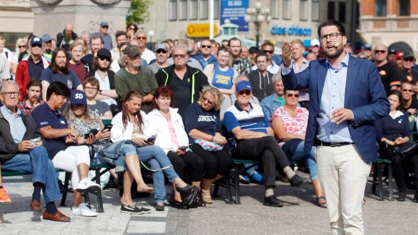 A man gestures in front of an audience. He is wearing a headset and is holding a political speech outdoors.